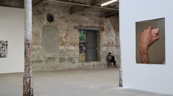 Fabrikhalle, in der Fotos aufgehängt sind. Auf dem Foto im Vordergrund rechts sind zwei Hände zu erkennen. Links eine rostige Säule. Im Hintergrund sitzt eine MItarbeiterein der Galerie auf einem Stuhl.