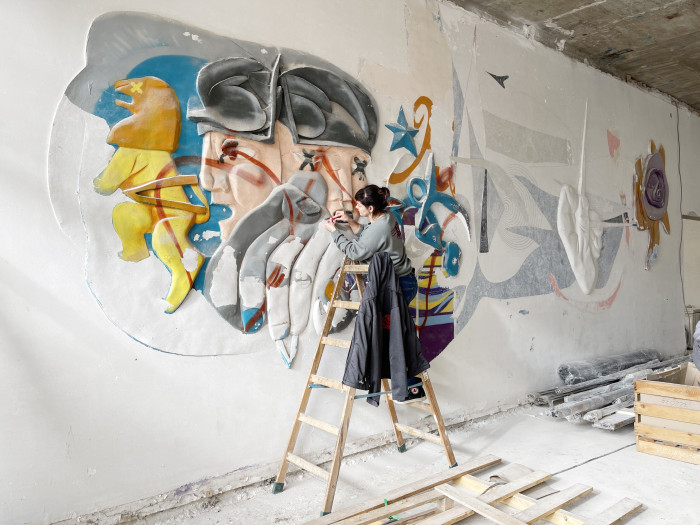 Frau steht auf Leiter und restauriert ein Wandgemälde. Ort: Gästehaus am Park Leipzig, Künstler: Bernhard Heissig